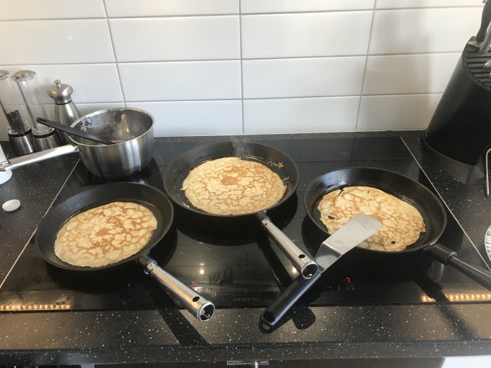 Bredd häll med tre stekpannor med pannkakor på en svart spishäll, kastrull och kryddkvarnar i bakgrunden.