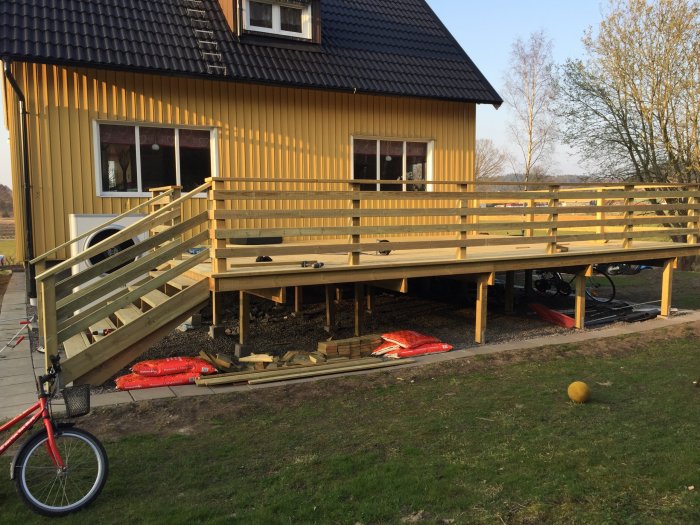 Gul trähus, stor veranda under konstruktion, trädgårdsredskap, cykel, soligt väder, gräs, utanför.