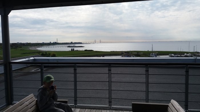 Barn sitter på bänk och tittar på havsutsikt med bro i bakgrunden.