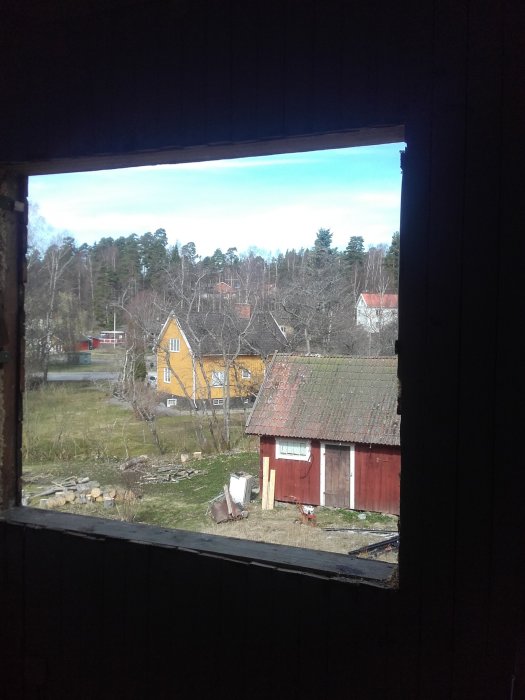 Utsikt genom en öppning utan fönster i en byggnad, visar en trädgård och röda samt gula hus.