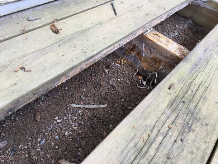 Ruttnande trädäck med synliga reglar och fuktig underlag av grus och sand.