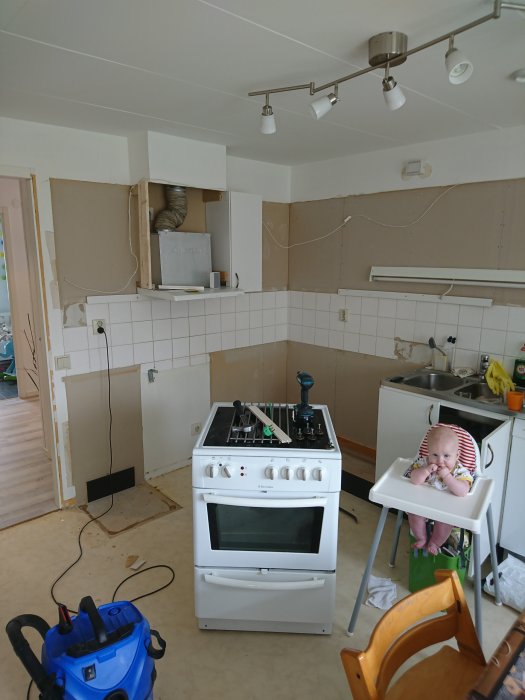Ett kök under renovering med borttagna skåp och en spis framflyttad, samt ett barn som tittar på från en barnstol.