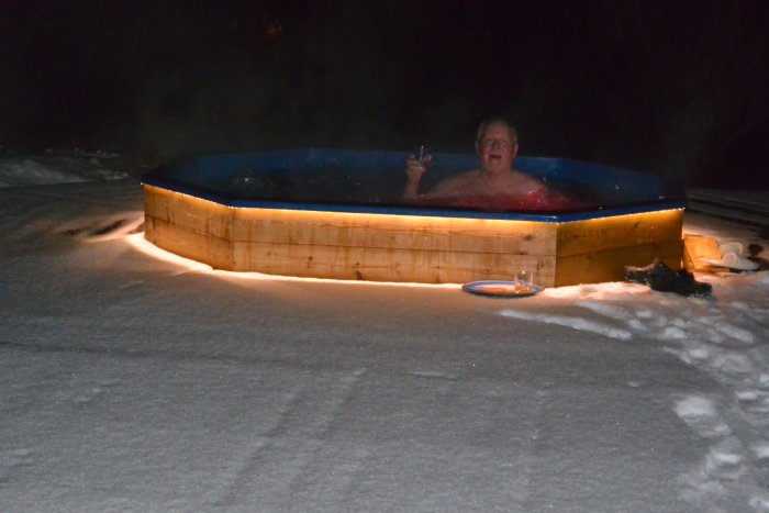 Person i en uppvärmd utomhusbadtunna omgiven av snö med ett glas whisky.