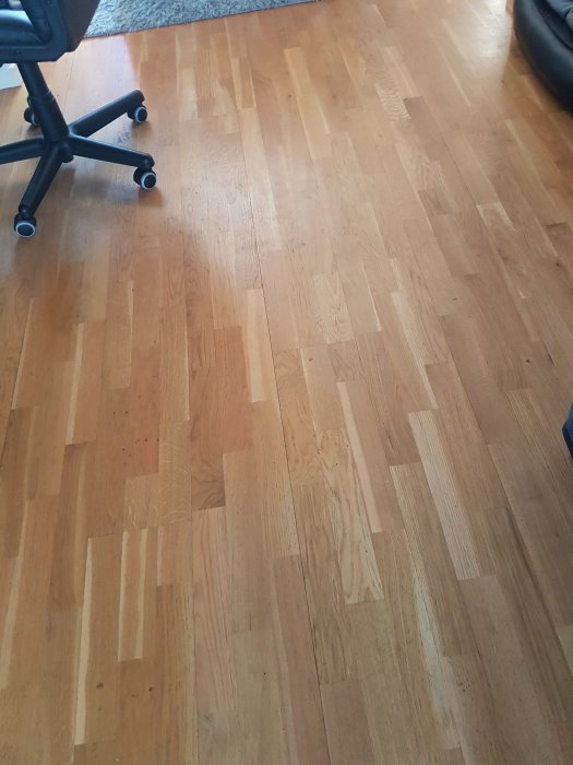 alt="Golv i vardagsrum med ljust träparkett möjligen från 60-talet, delvis täckt av en svart kontorsstol till vänster.