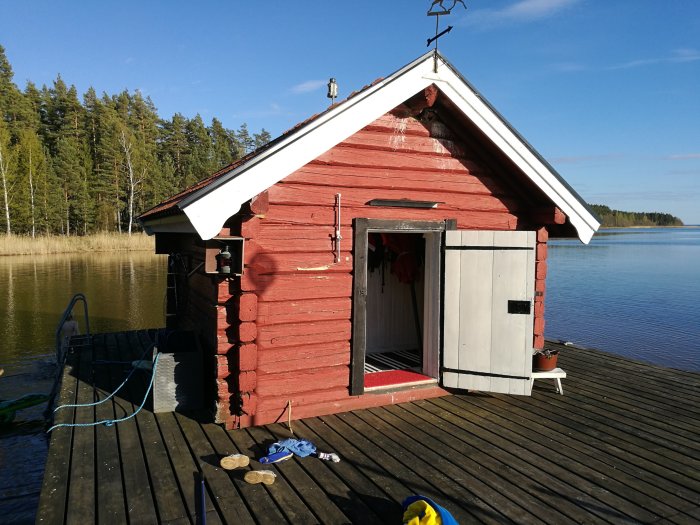 Röd timmerstuga med vit dörr på en brygga vid en sjö, skador på stockarna syns ej tydligt.