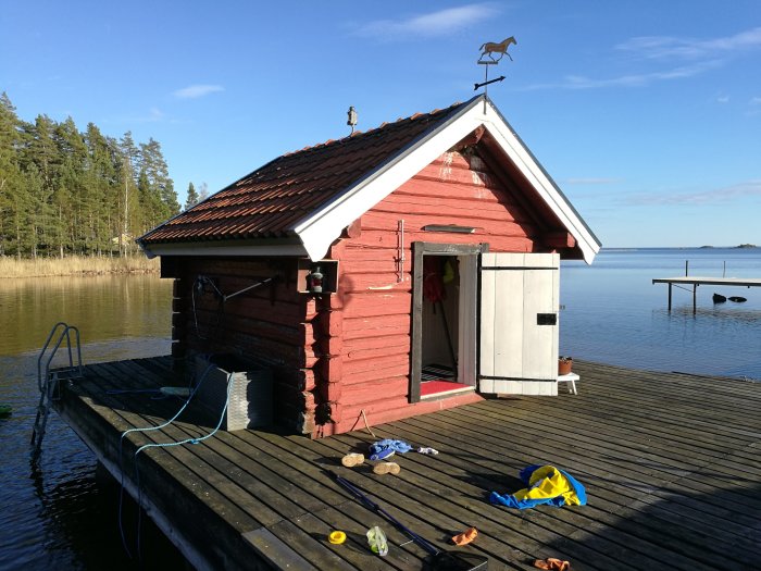 Rött timmerhus med vita knutar beläget på en brygga vid vattnet, skador ej synliga i bilden.