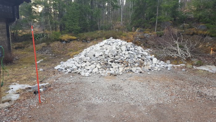 Hög med gråa byggstenar på en grusplan med skog och gräsmark i bakgrunden samt en röd mätpinne till vänster.