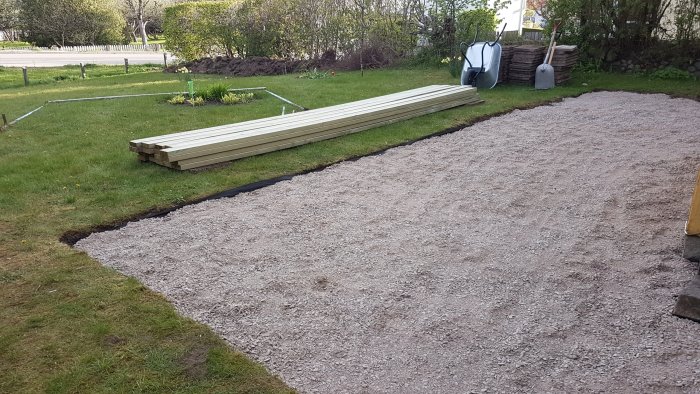 Trädgårdsarbetsområde med förberett grusunderlag, byggmaterial, skottkärra och trädgårdsverktyg.
