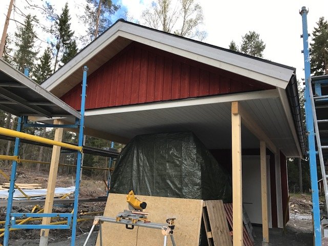 Carport under konstruktion med synliga takbjälkar, byggmaterial och geringssåg i förgrunden.