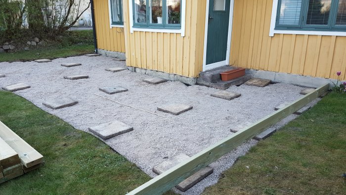 Förberedd trädgård för trallbygge med grusbas och utspridda betongplattor framför ett gult hus med gröna fönsterkarmar.