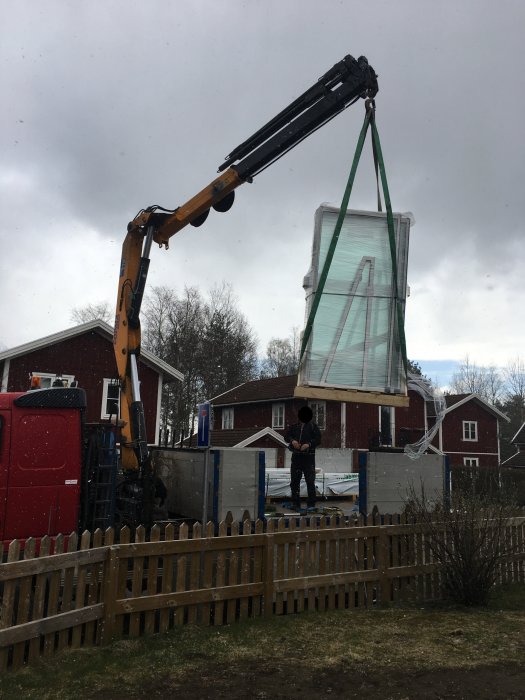 Kran som lyfter en förpackad glasparti till ett uterum över ett staket med man som observerar.