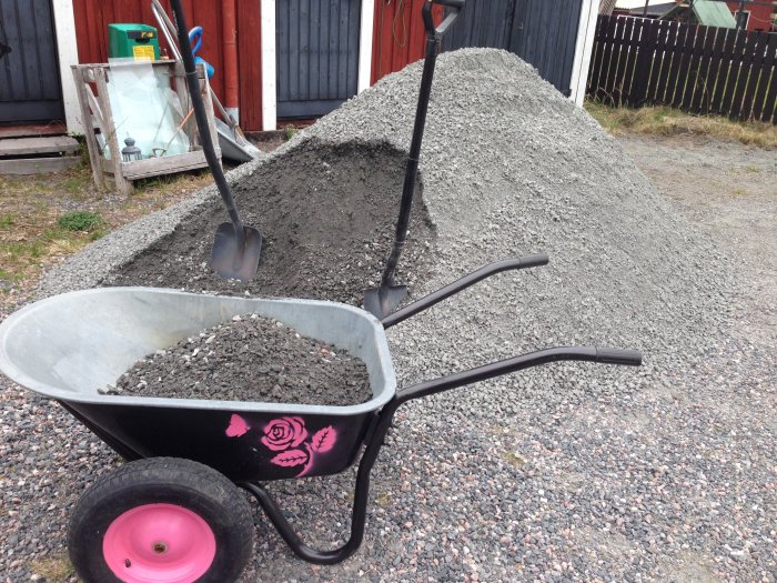 En skottkärra med rosa hjul och dekorationer framför en stor hög med grus, bredvid en spade.