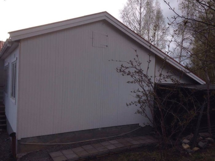 Vit husfasad med nylagd svart vindskiva vid taket och nyrenoverat trädgårdsförråd intill garaget.