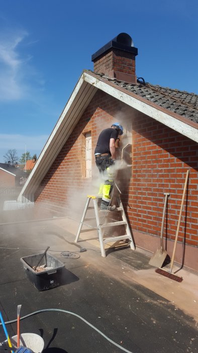 Person som sågar hål för fönsterinstallation i tegelhusets fasad, stående på en stege.