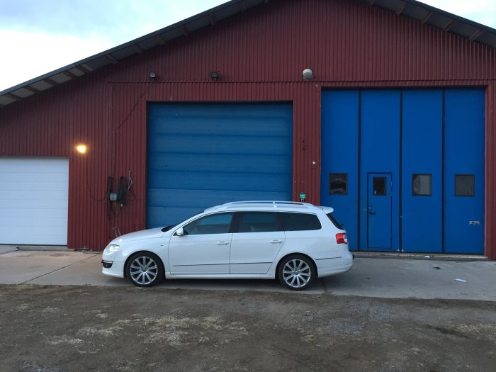 Vit bil framför stort garage med blå dörrar, däckbyte utförs utomhus, rödfärgad byggnad.