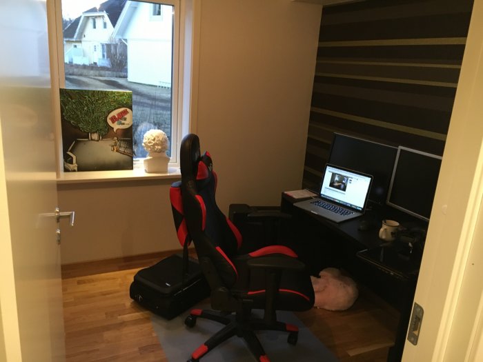 Hemmakontor med svart-röd gamingstol, skrivbord med laptop och dubbla skärmar, och randig tapet.