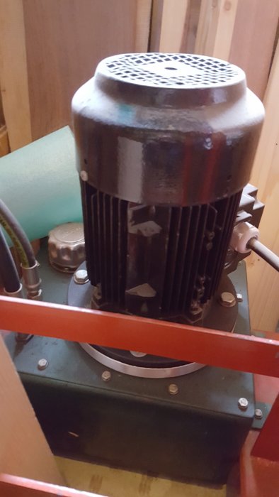 Hembyggd press med svart motor och röda metallramar, placerad i ett utrymme med träväggar.