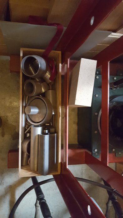 Delar av en handbyggd press med metallkomponenter och röda stålramar, sedd i en verkstadsmiljö.