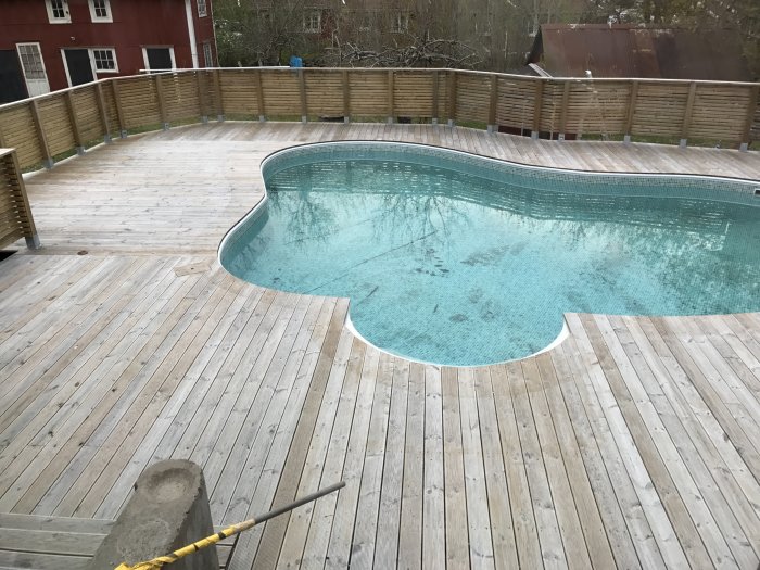 Nyinstallerat staket och trall runt en inbyggd pool med vatten, väntar på oljebehandling, under en mulen himmel.