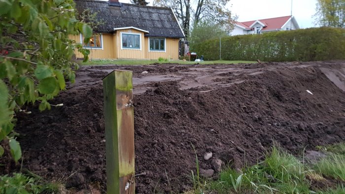 Nyplanerad jord framför ett gult hus med grön häck i bakgrunden, markeringsstolpe i förgrunden.