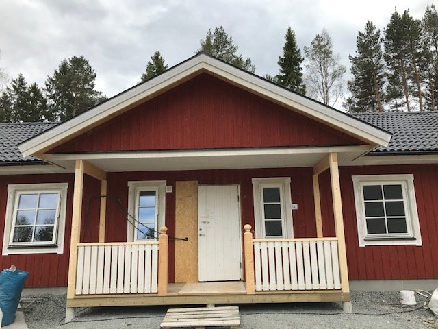 Rödmålat hus med nymålad panel och temporär dörr, räcke på veranda och skog i bakgrunden.