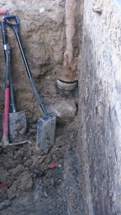 Dräneringsgrävning vid ett 60-tals hus där en stor sten syns under ett stuprör, med spadar i förgrunden.