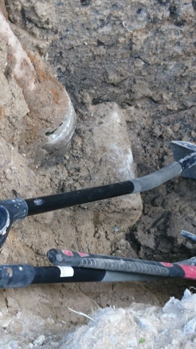 Grävd grop visar del av stuprör och en stor betongplatta nedanför, omgiven av jord och grus, verktyg syns också.