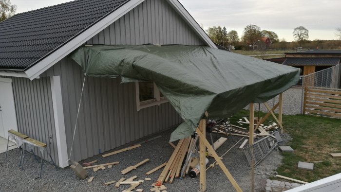 Delvis uppbyggd carportstomme täckt med presenning vid ett grått hus.