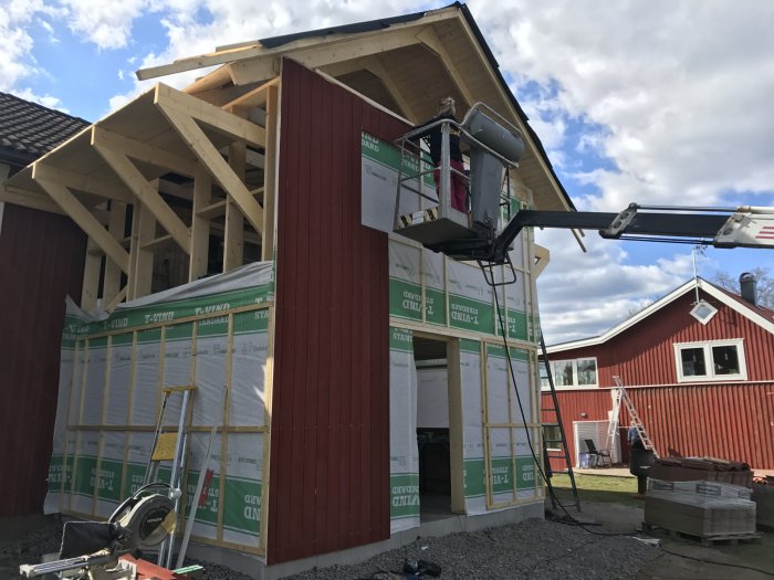 Byggarbetsplats med halvfärdigt trähus, vindskyddsisolering och en skylift vid sidan.