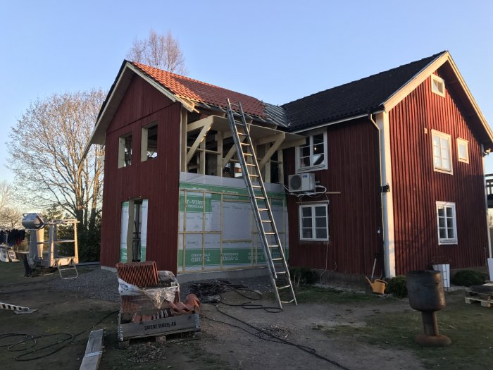 Byggplats med rött hus under renovering, delvis ställning, tegeltak i kvällsljus.