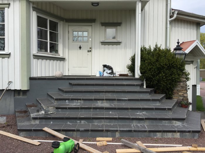 Slitna entretrappor i sten framför hus med vit dörr och fönster, renoveringsmaterial syns på marken.