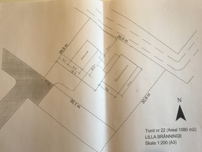 Skiss av en tomt med måttangivelser där ett hus och garage är placerade, med kompassriktning norr markerad.