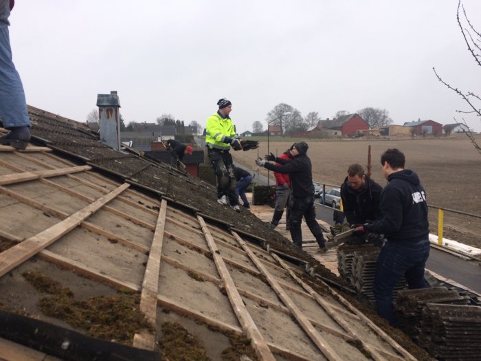 Arbetare som demonterar gammal takbeläggning på ett hus med landsbygd i bakgrunden.