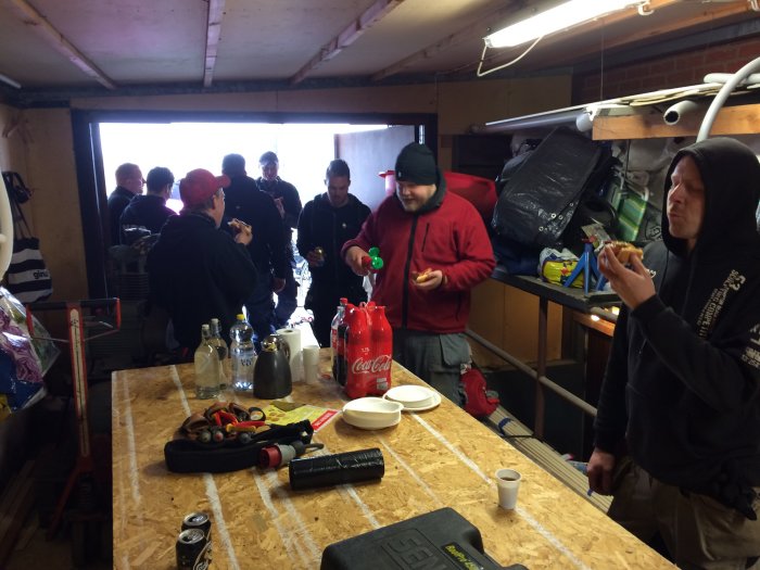 Grupp människor i verkstad samlade kring bord med snacks och dryck, arbetsverktyg synliga.