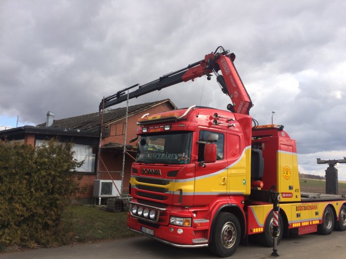 Röd Scania bärgningsbil med kran parkerad framför ett hus under molnig himmel.