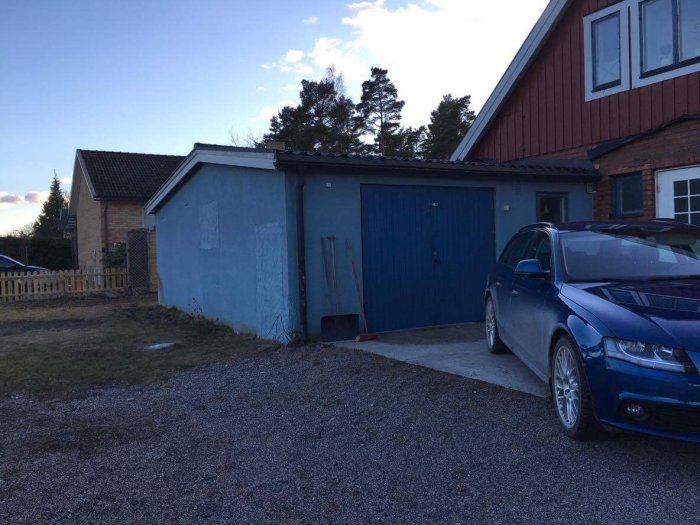 Blått garage intill rött hus med ojämnt målad vägg och murade delar, grusuppfart och blå bil framför.