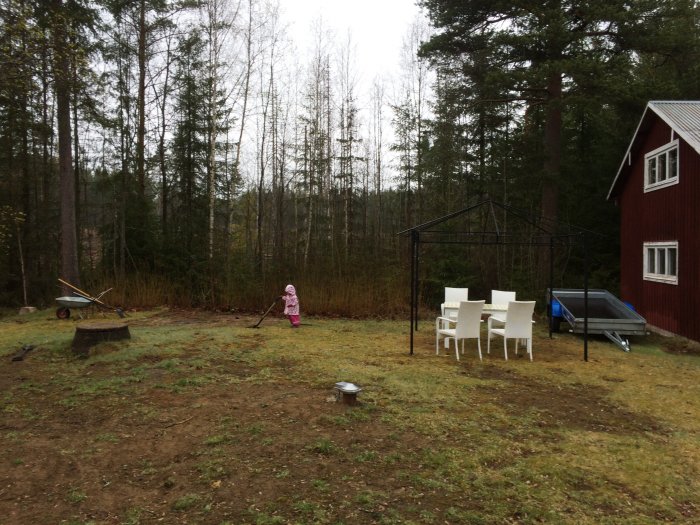 Trädgårdsyta med rödfärgat hus, en paviljongstomme, vita plaststolar och en barnlekplats, planeras för däckbygge.