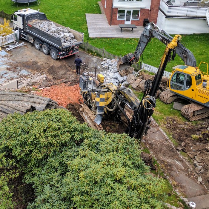 Lastbil som levererar grus och en grävmaskin arbetar i ett bostadsområdes trädgård.