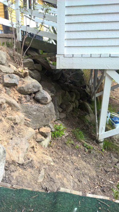 Lutande tomt med en oregelbunden naturstenmur under ett hus, vissa stenar verkar lösa.