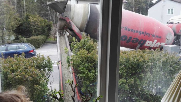 Betongbil som gjuter betong sett genom ett fönster, med gröna häckar i förgrunden.