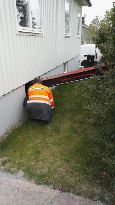 Arbetare i orange och grå arbetskläder böjer sig för att jobba under ett husfundament, omgiven av grönt gräs.