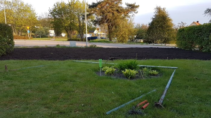 Nyjordad trädgård med spaljé och krattning, gräsmatta i förgrunden och blomlåda med växter.