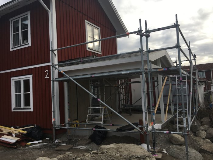 Byggställningar framför rödfärgad husfasad under renovering med synliga byggmaterial och rör på marken.