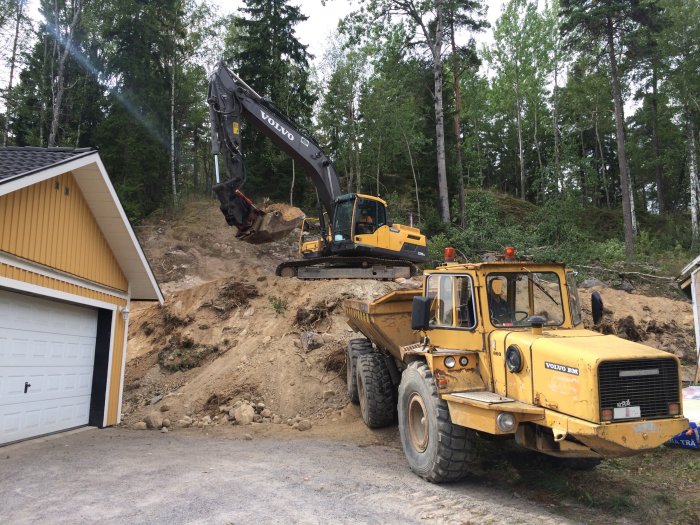 En grävmaskin och en dumper vid markarbeten nära en gul villa.