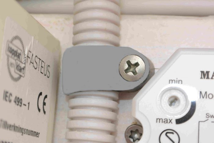 Grå elektrisk klammer monterad på vit rörformad kabel med CE-märkning synlig.