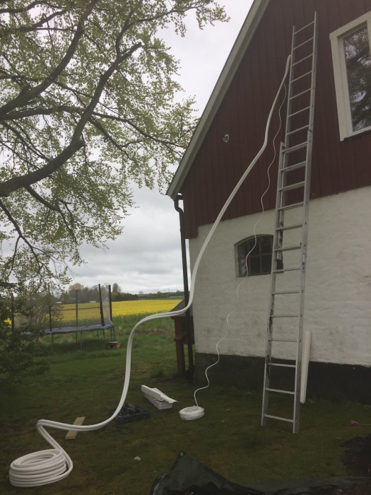 Nyinstallerad värmepump med rör som sträcker sig längs en röd lada med vit knut, stege och trädgård.