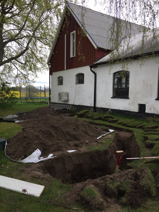 Grävd trädgård med öppna diken nära en rödvit lada för installation av värmepump och avloppsrör.