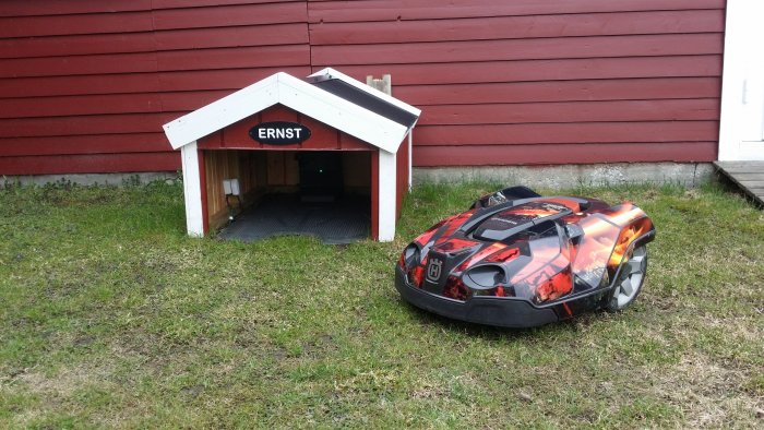 Vit hundkoja med namnet "ERNST" och en robotgräsklippare med flamdesign framför röd husvägg.