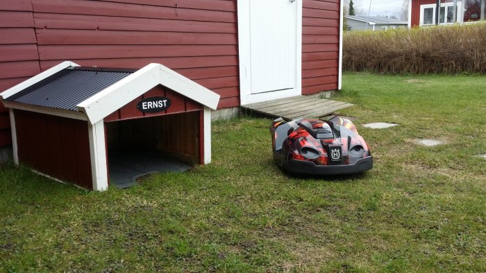 Hundkoja med namnet "ERNST" och en leksaksbil vid gräsmatta intill rött hus.