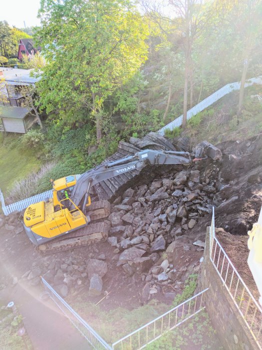 Grävmaskin märkt Volvo arbetar bland stora stenblock på en byggarbetsplats med grönska runt omkring.